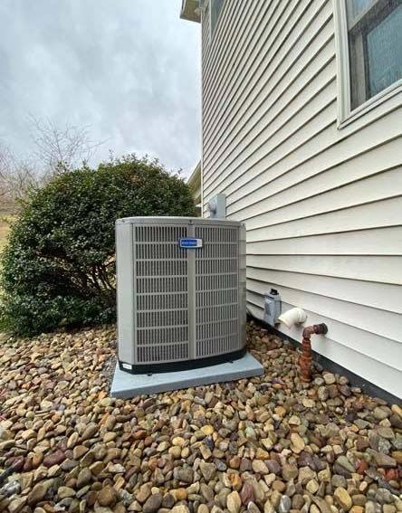 An outdoor HVAC condenser unit sits on a concrete pad next to a beige-sided house, surrounded by decorative landscape rocks.