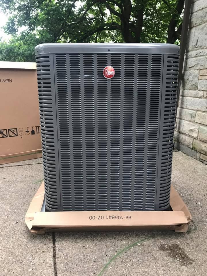 A new gray Rheem central air conditioning unit sitting on a cardboard base outdoors on a concrete patio.