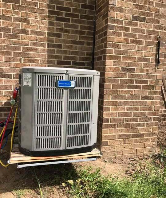 An outdoor air conditioning unit sits on a platform attached to a brick wall, with colorful gauge hoses connected.