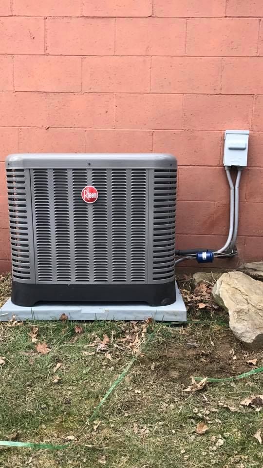 A Rheem HVAC unit sits on a concrete pad against a red brick wall next to an outdoor electrical disconnect box.