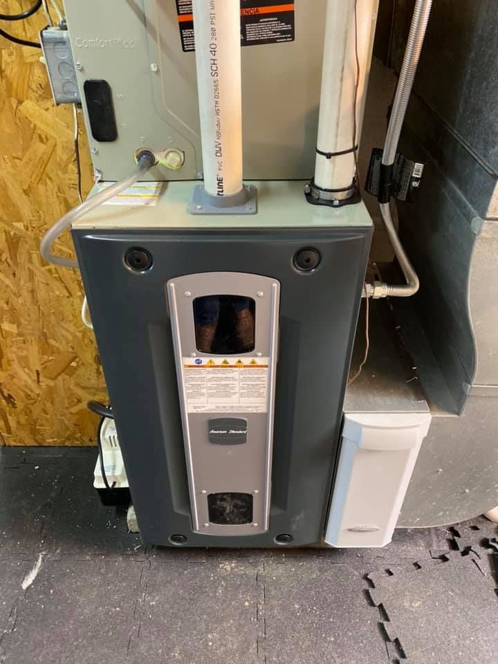 A gray, upright HVAC furnace unit with white PVC pipes attached, situated on a dark mat against a wooden wall.