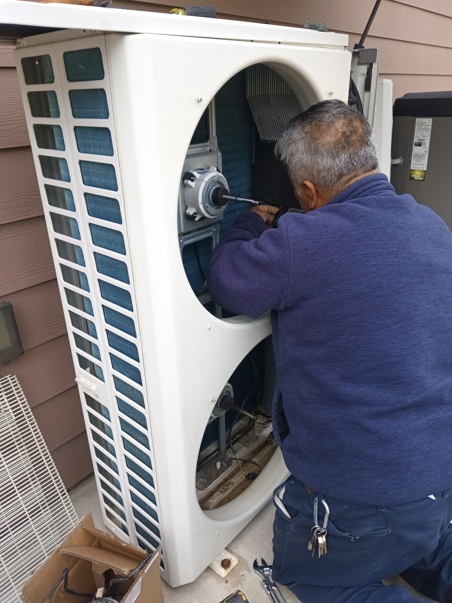 Man repairs an outdoor air conditioning unit; he wears a blue sweater and is using tools.