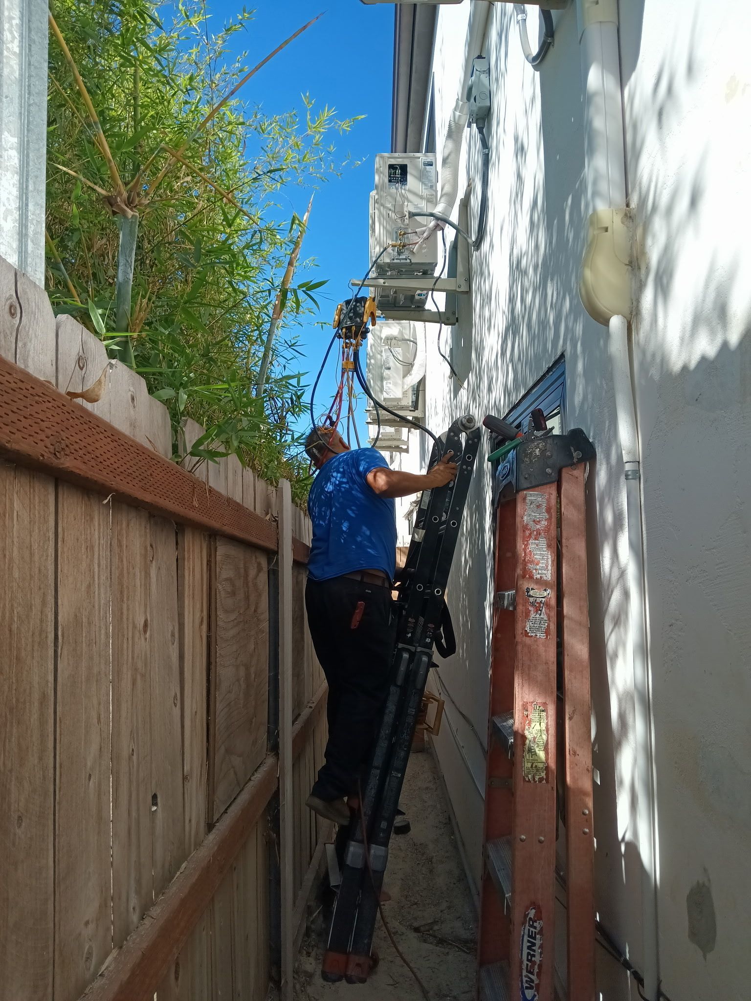 Person on ladder working on AC unit mounted on a white wall, narrow space between fence and building.