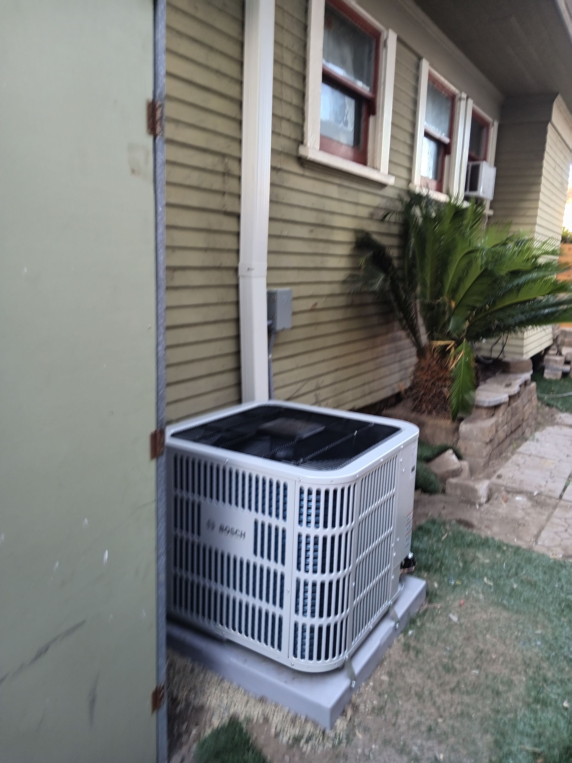 Air conditioning unit outside a green house next to a door and a plant.