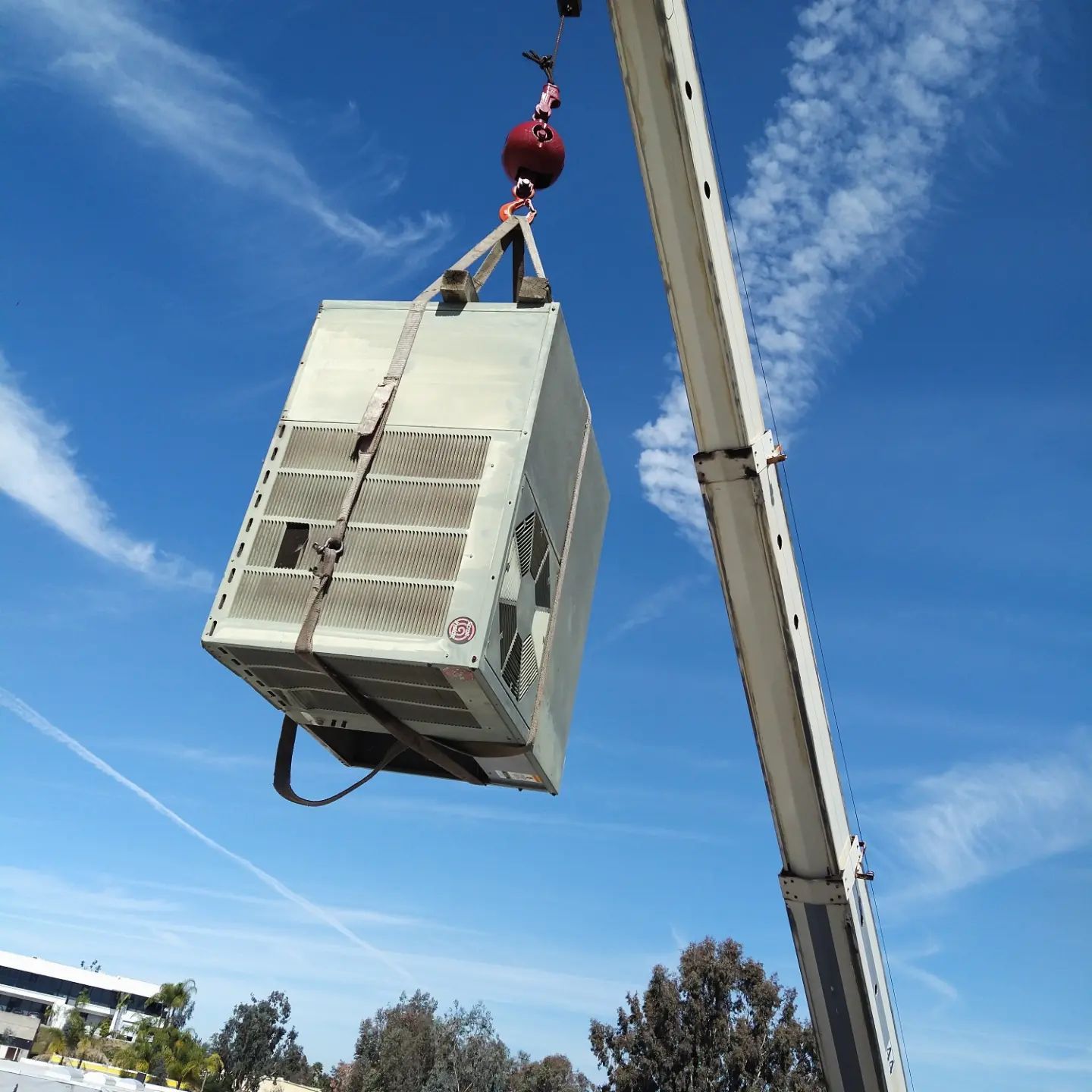 A large metal unit, strapped, is lifted by a crane against a blue sky.