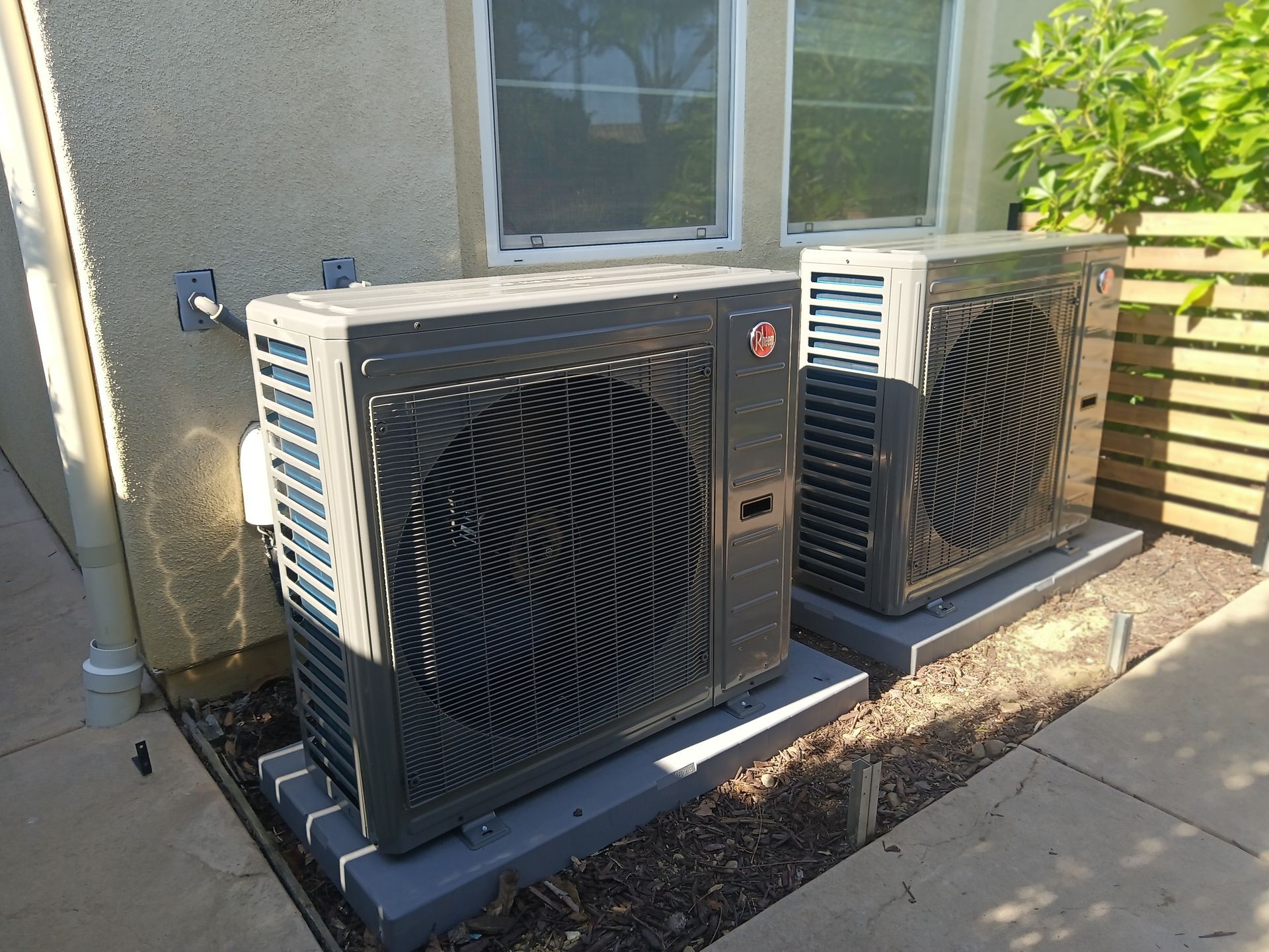 Two dark gray Rheem HVAC units outside against a stucco wall, on concrete pads.