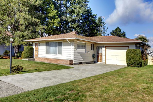 A one-story suburban house with cream-colored siding, a brown roof, and an attached garage under a blue sky.