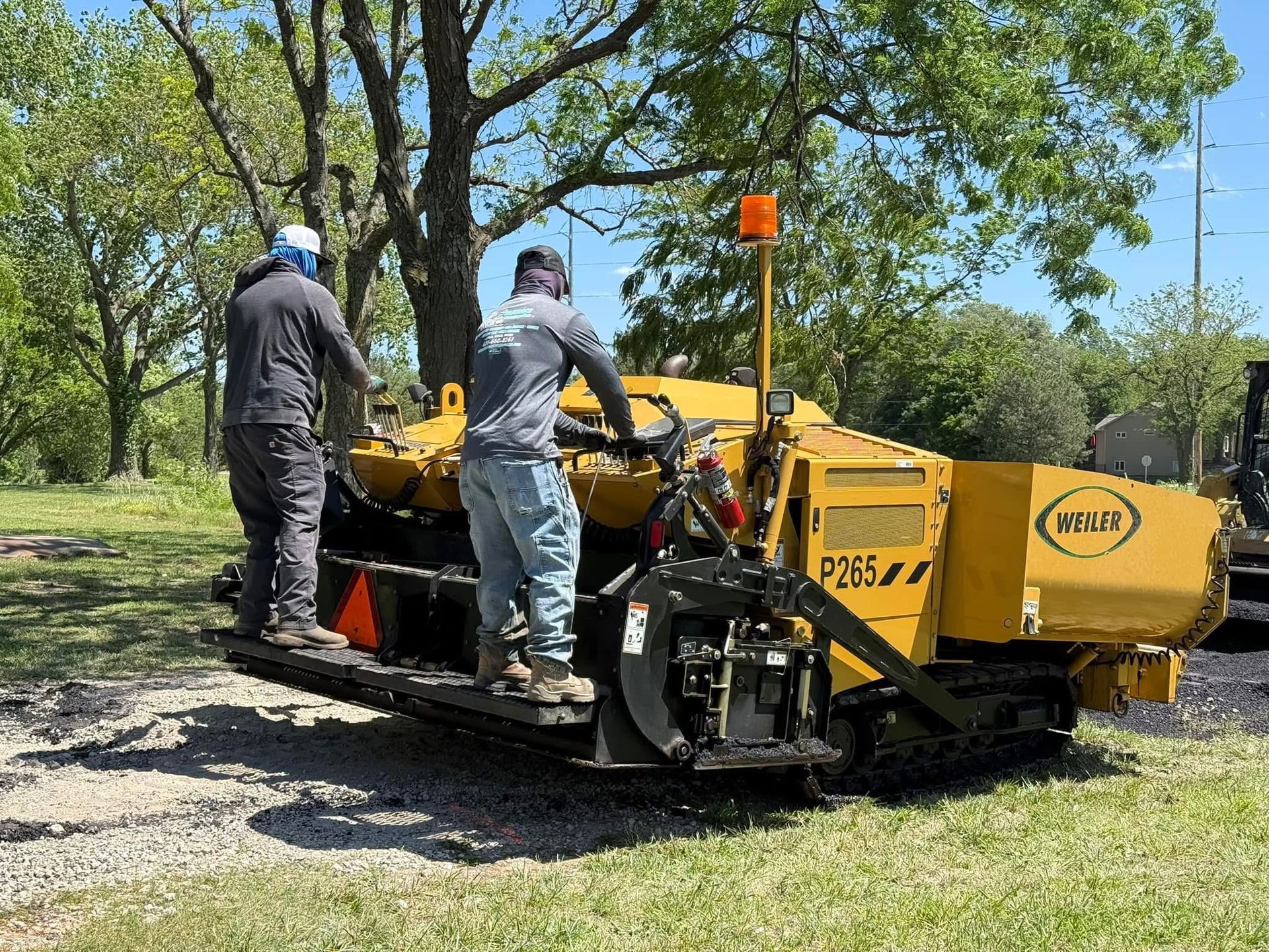 Two workers operate a yellow asphalt paver on a sunny day in a grassy, wooded area.