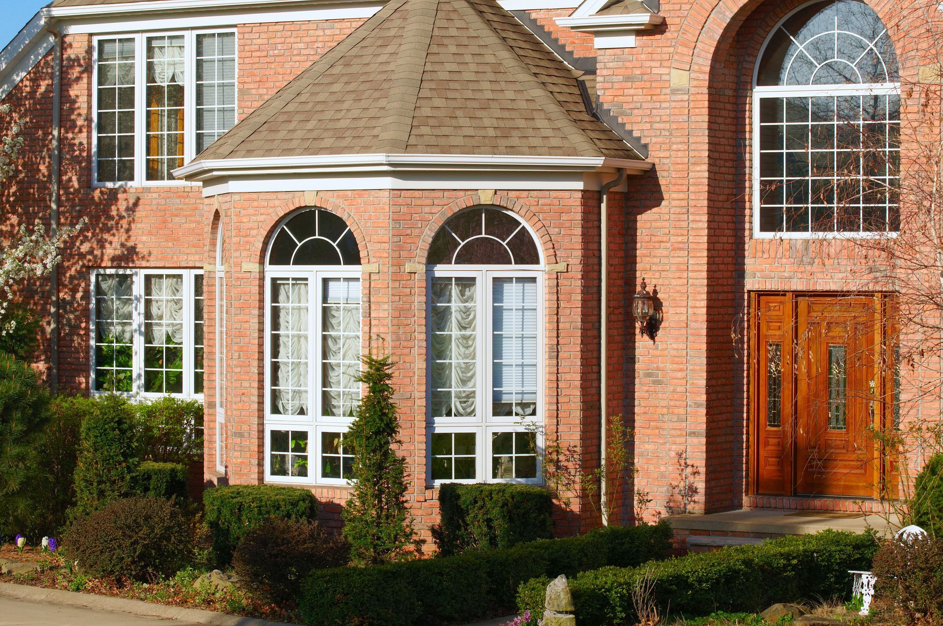 Brick suburban house with arched windows, white trim, and a wooden front door surrounded by shrubs.
