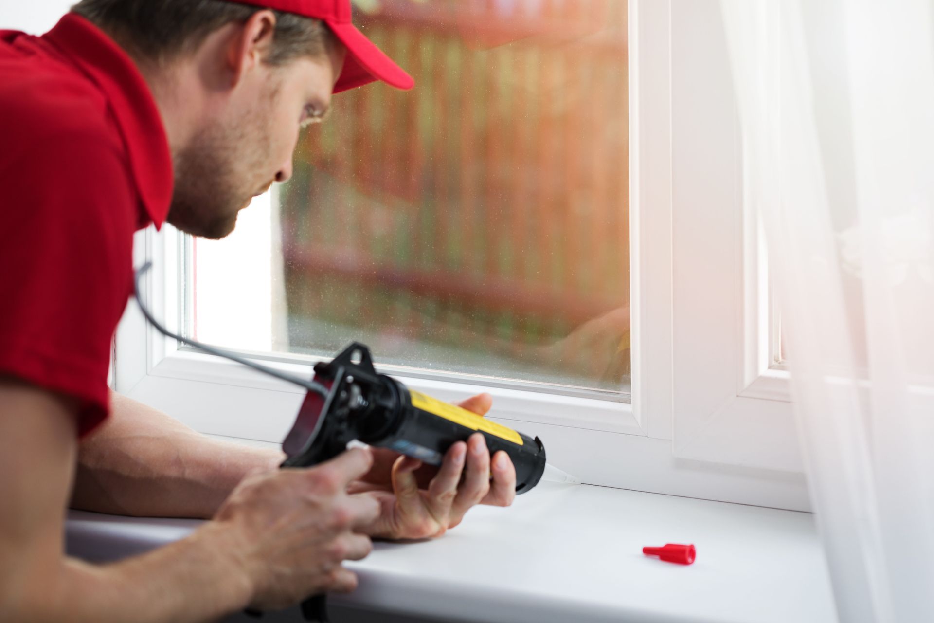 Worker applying sealant to a white window frame with a caulking gun.