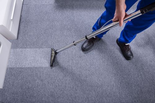 Person vacuuming a gray carpet with a long silver vacuum cleaner in a hallway