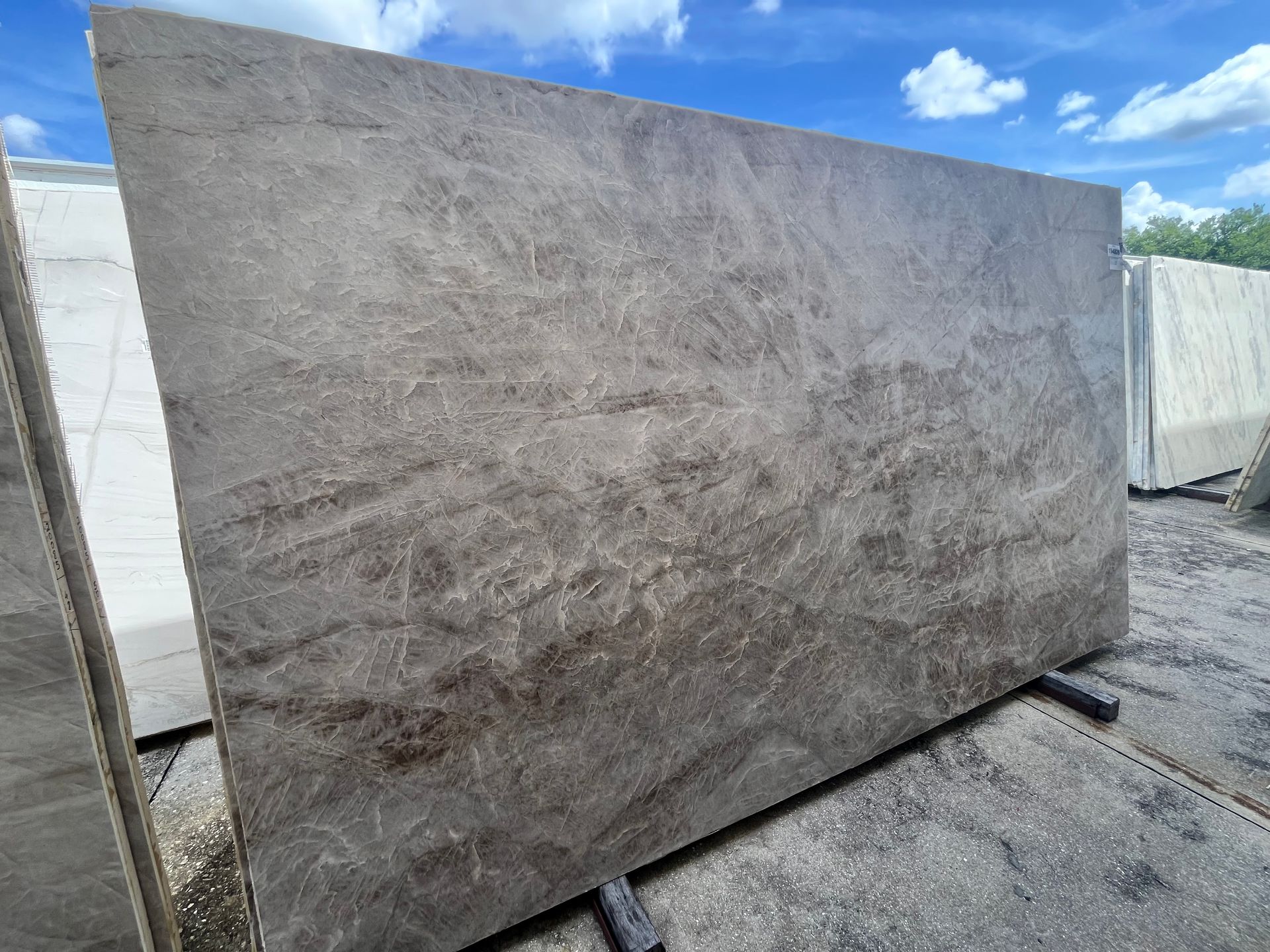 A large, polished slab of grey marble with subtle brown veining, standing upright in an outdoor stone yard.