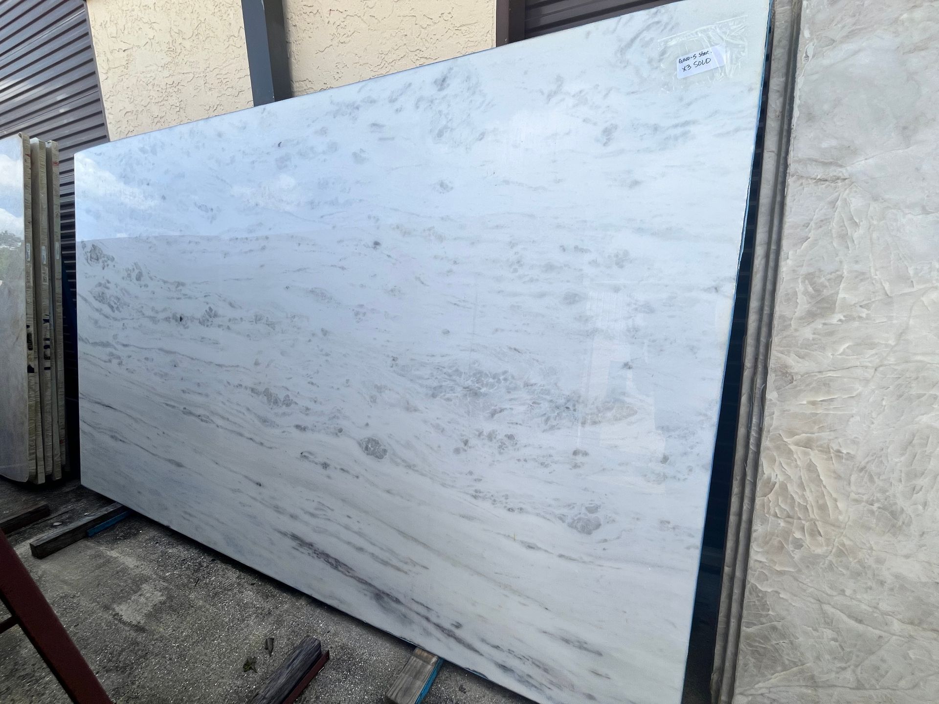 A large, polished slab of white marble with subtle grey veining displayed at an outdoor stone yard.