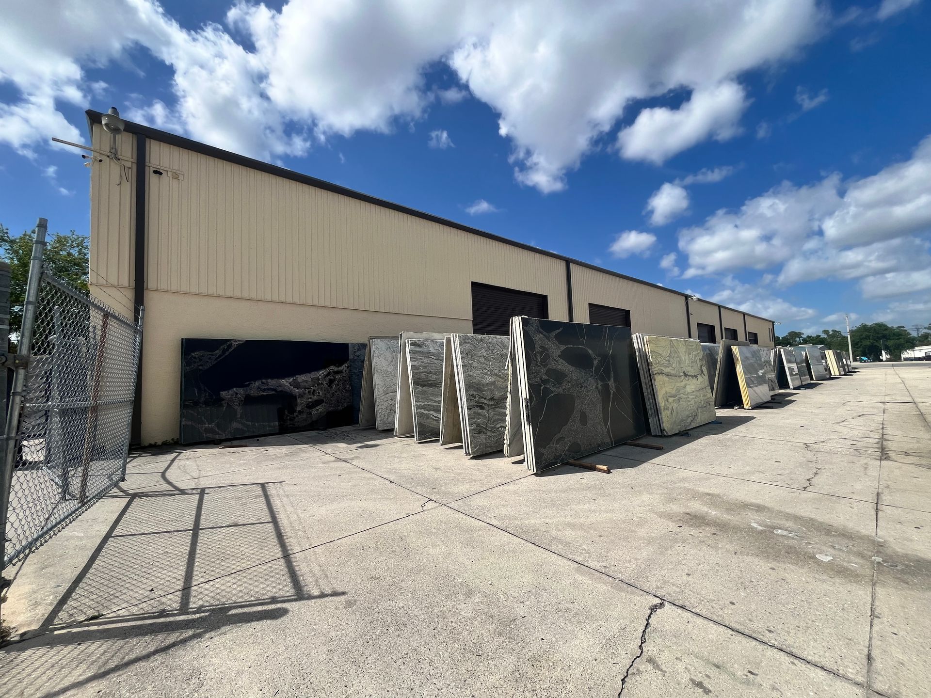 Large stone slabs of various colors and patterns stored outside against the side of a warehouse building under a blue sky.