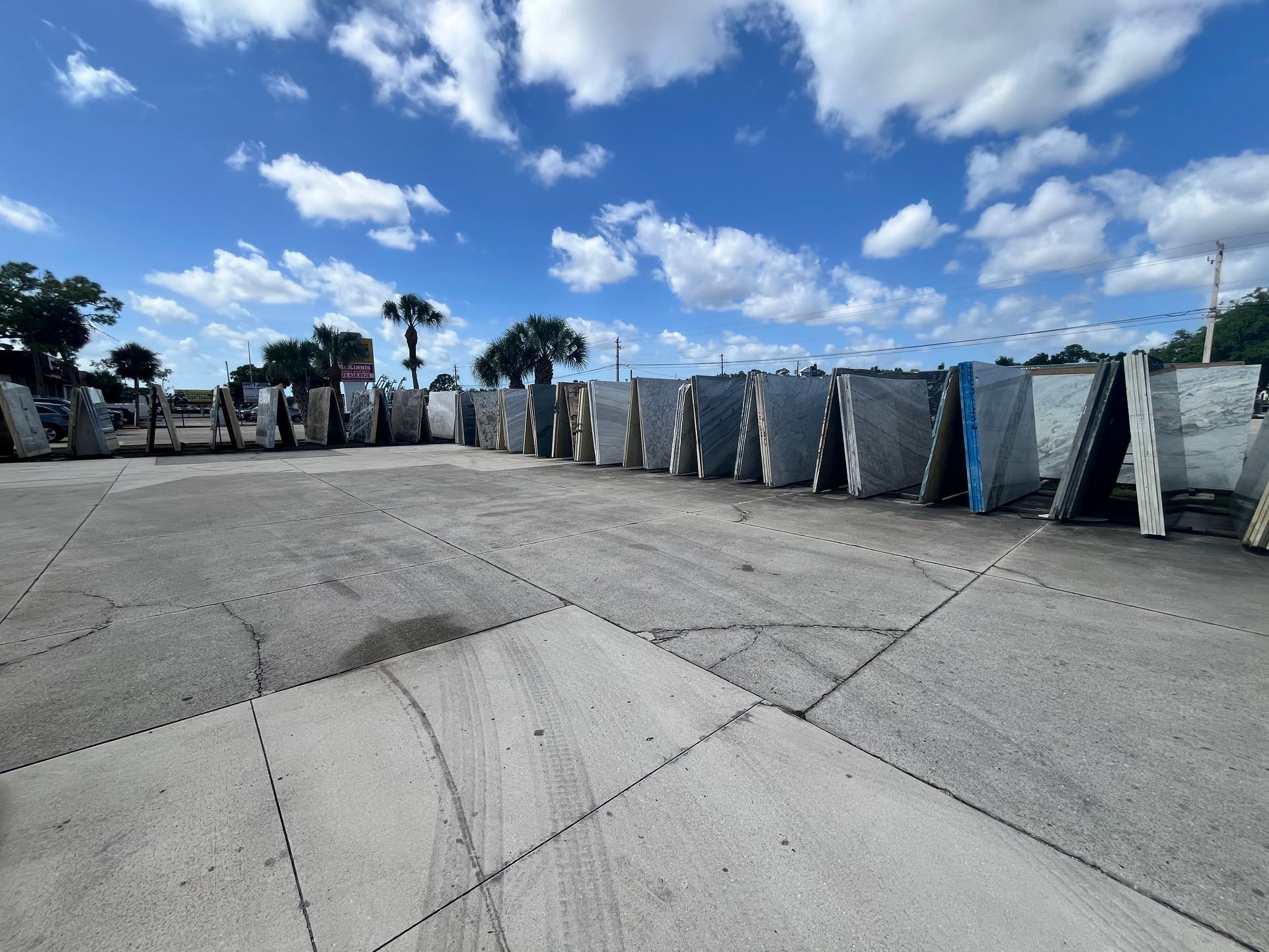 Large stone slabs displayed in long rows on an outdoor concrete lot under a blue sky with scattered white clouds.