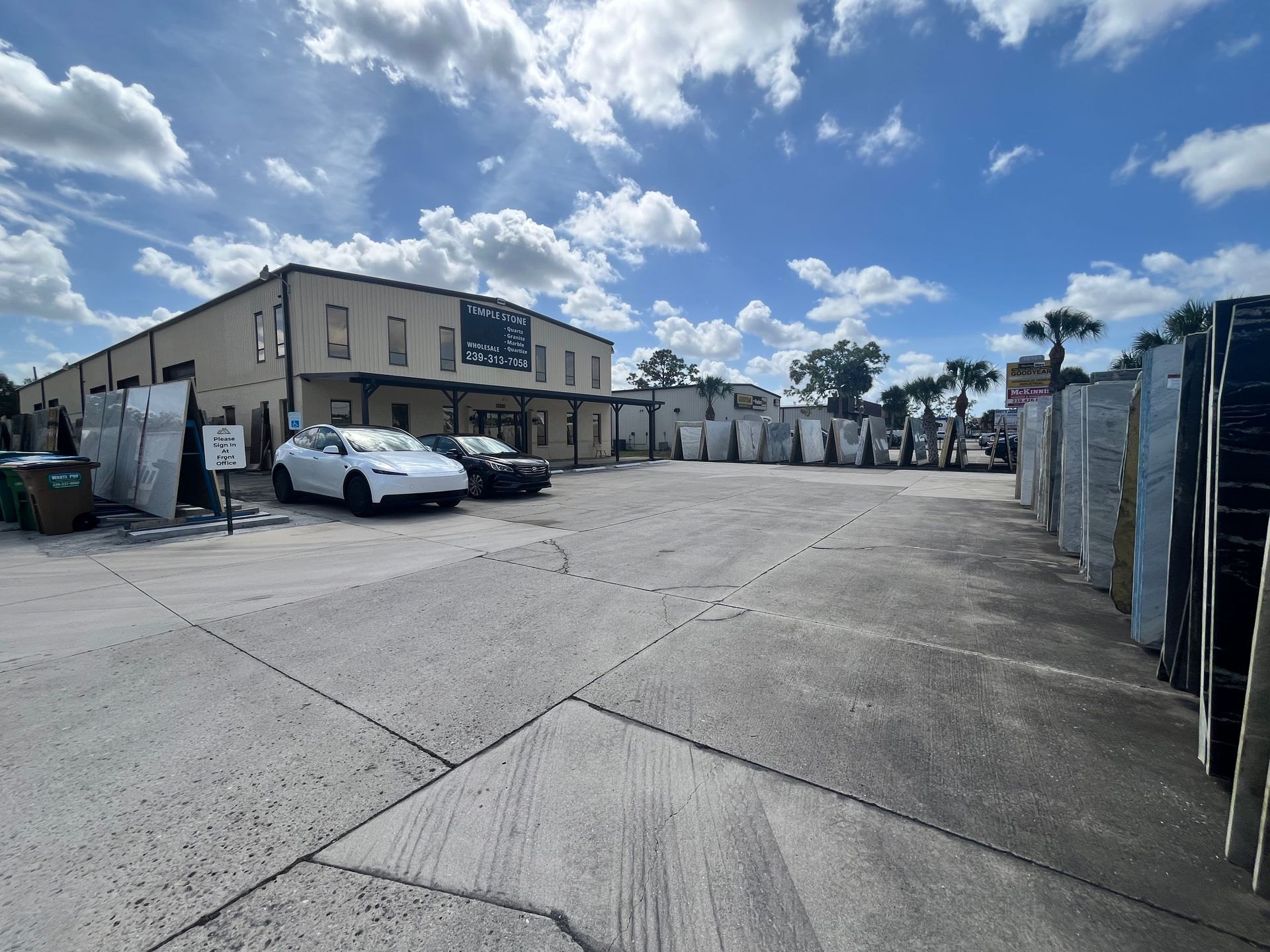 A light-colored industrial warehouse building with stone slabs stored outside in a paved lot under a blue sky.