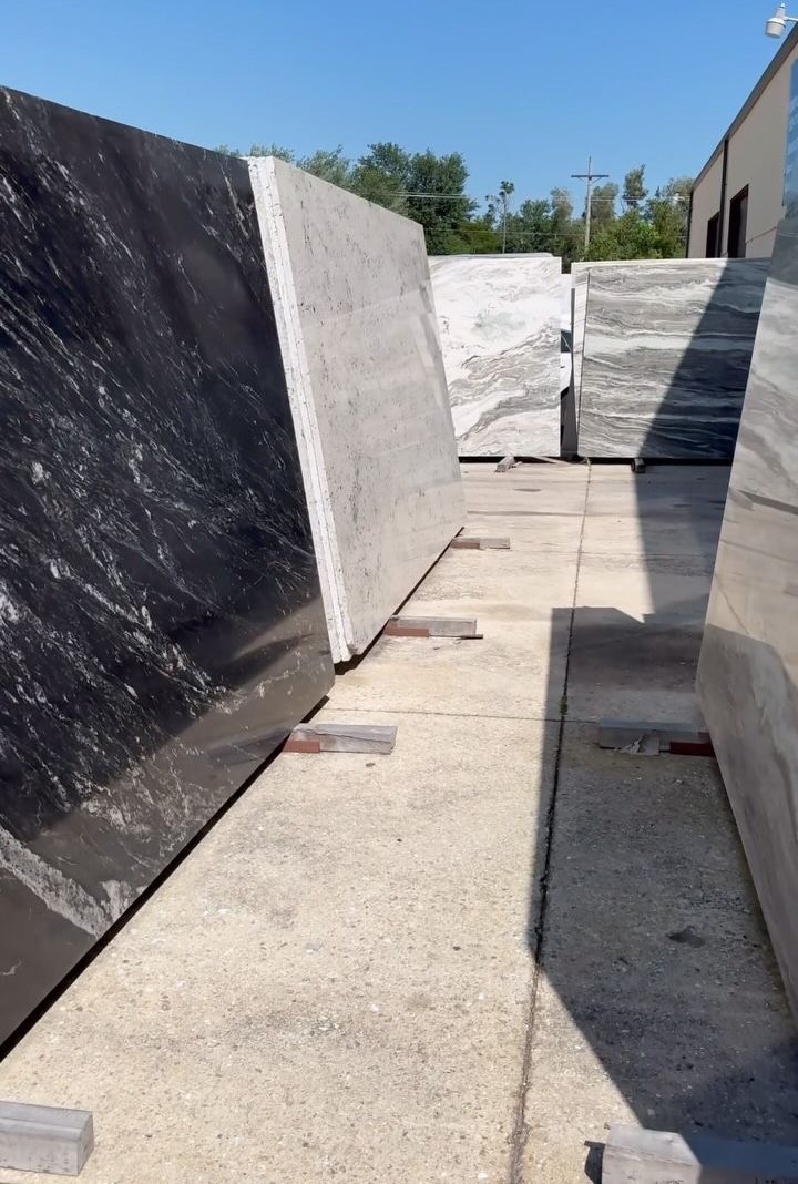 Rows of large, upright stone slabs of various colors and textures displayed in an outdoor yard under a clear blue sky.