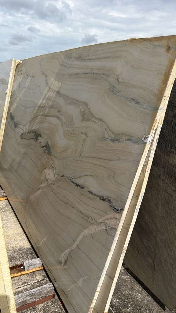 Large beige stone slab with wavy gray veining, standing outdoors in a yard with cloudy sky.