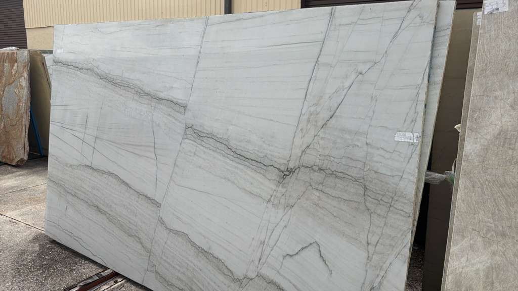 Large white stone slab with gray veining leaning outdoors against other slabs in a yard