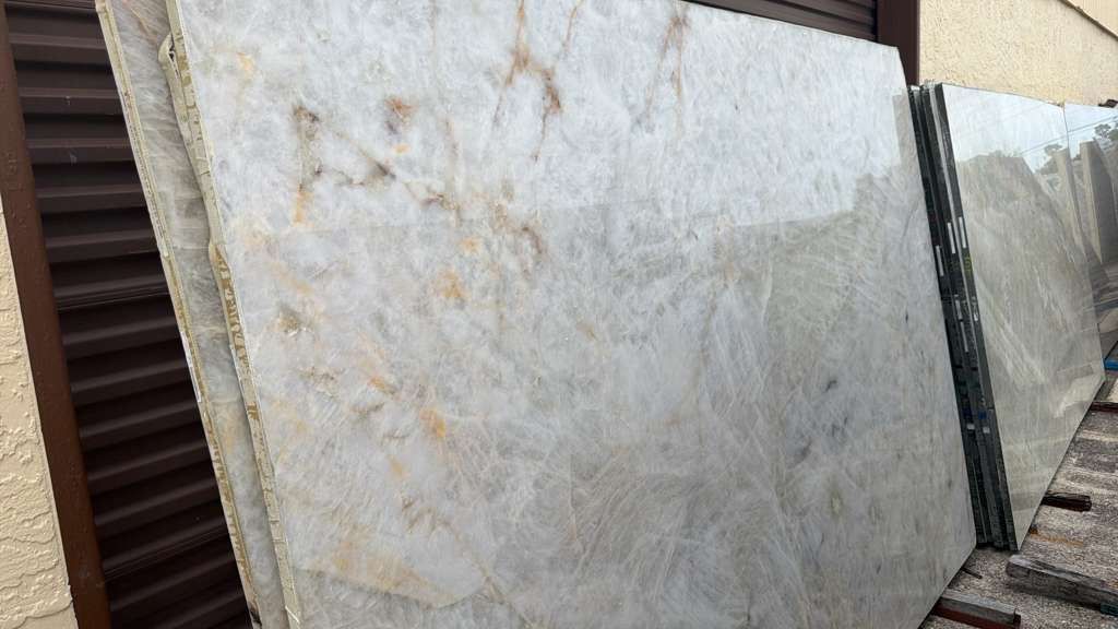 Stacked white marble slabs leaning outdoors beside a brown corrugated wall and metal fence.