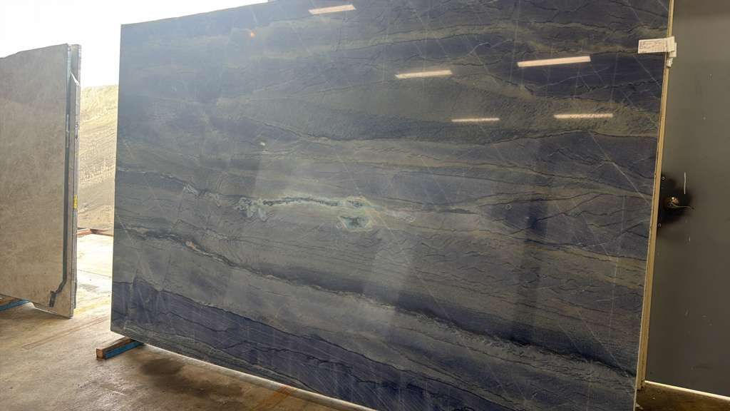 Large dark blue-black stone slab with horizontal veining, standing outdoors in a yard or workshop area.