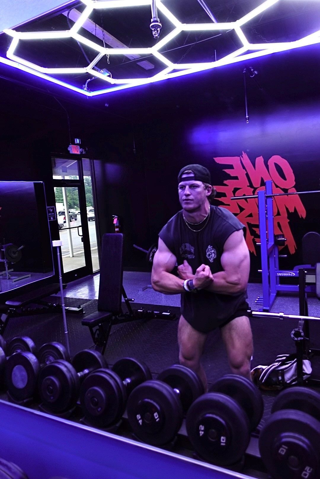 Jack Mulhern is standing in front of a row of dumbbells in a gym.