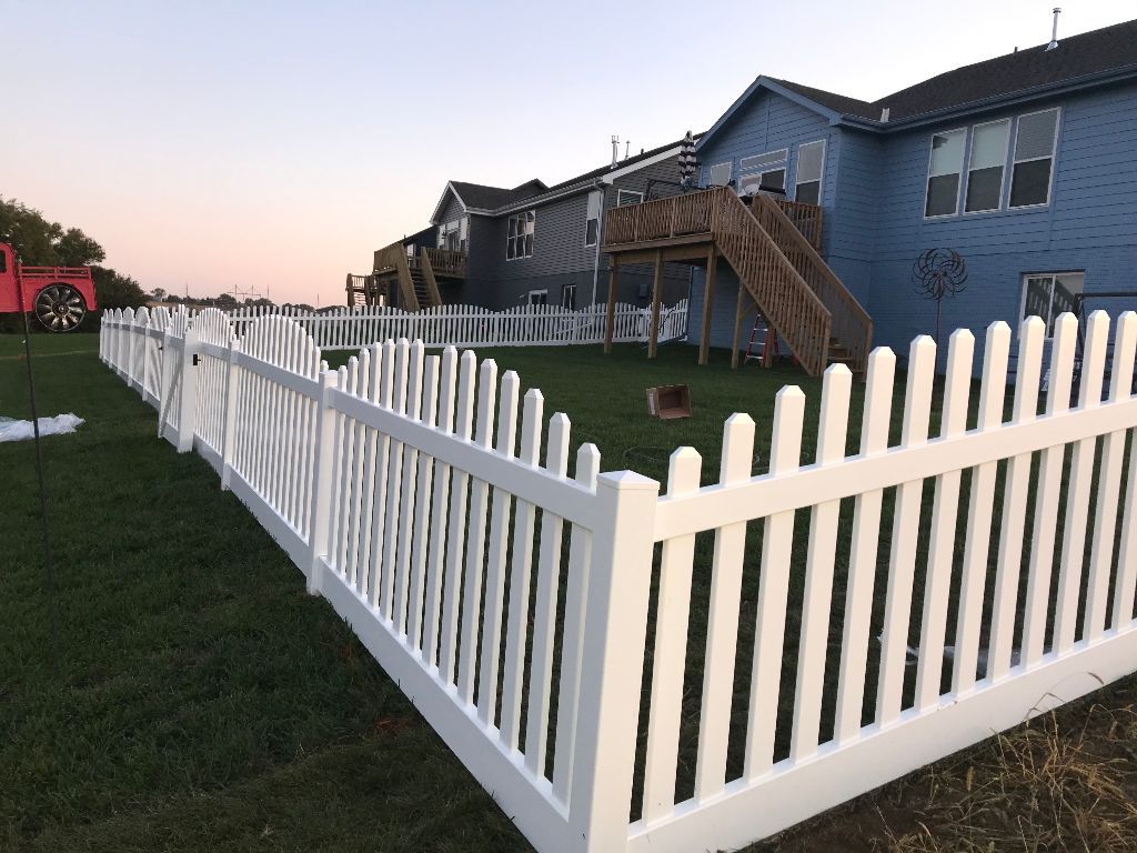A white picket fence surrounds a lush green yard in front of a blue house.
