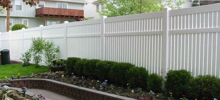 A white fence is surrounded by bushes and flowers in a backyard.
