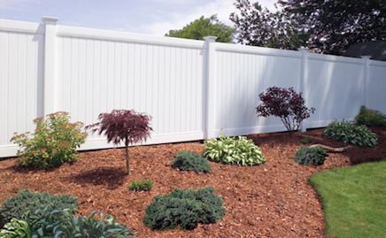 A white fence surrounds a garden filled with plants and mulch.