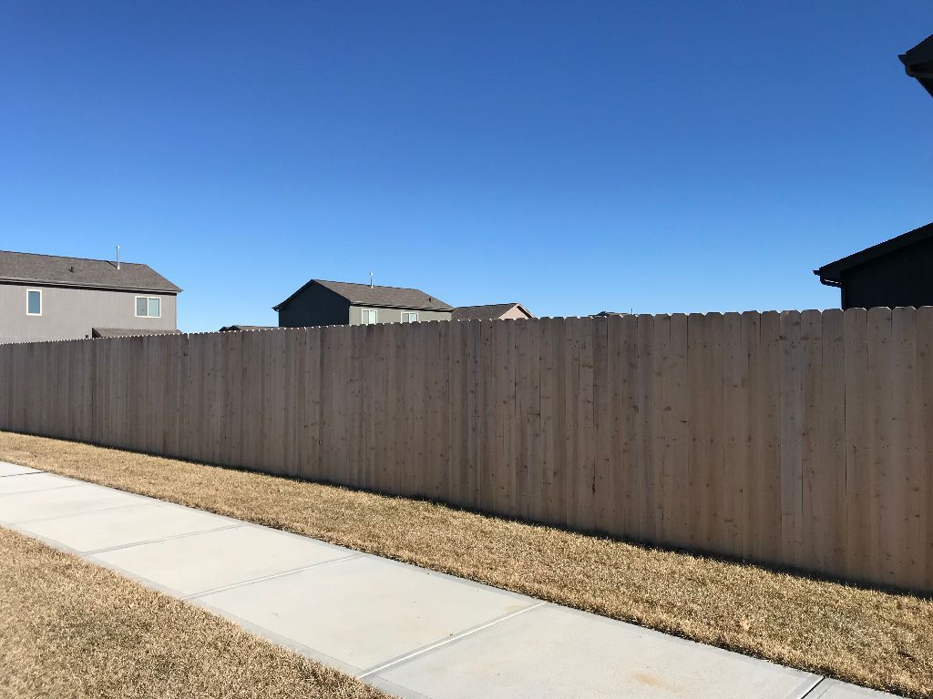 A wooden fence along a sidewalk with houses in the background.