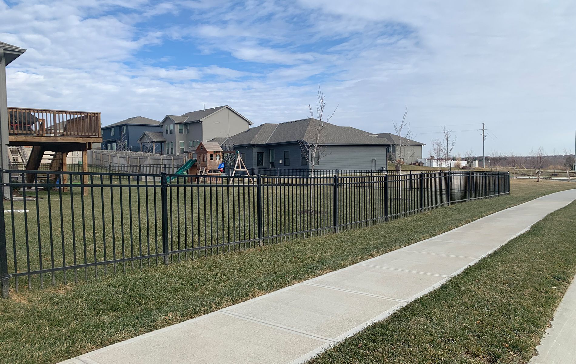 A sidewalk leading to a fenced in yard with houses in the background.