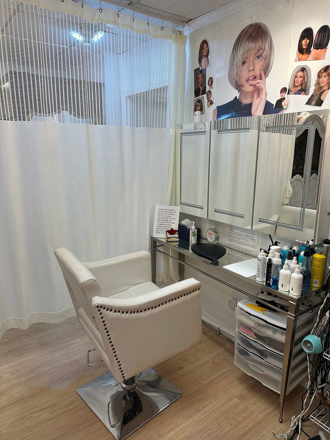 Salon chair beside mirrored styling station with drawers, posters, and white curtains in a bright room.