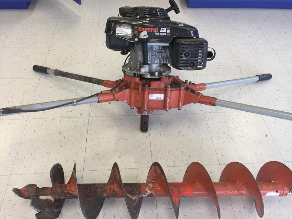 Gas-powered auger with a red drill bit on a floor. The auger has a black engine, red gearbox, and four gray handles.