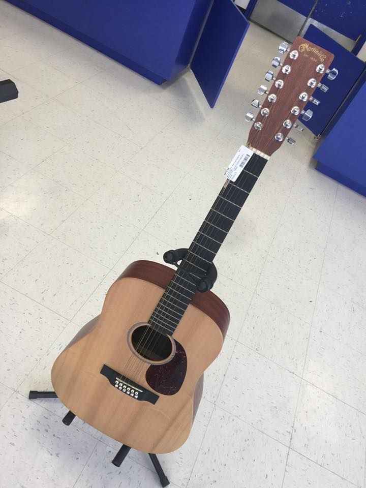12-string acoustic guitar on a black stand; beige wood, brown neck, and black fretboard.
