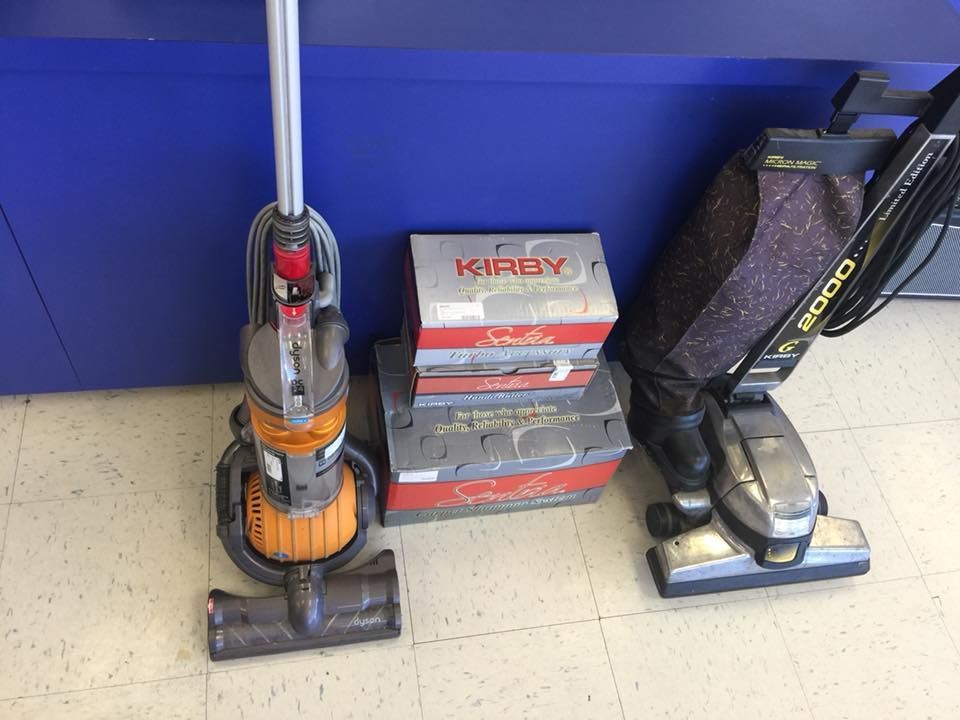 Dyson and Kirby vacuums with Kirby filter boxes against a blue wall on a tiled floor.
