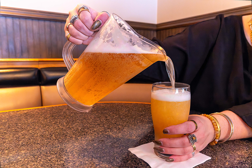 Hands pouring beer from a pitcher into a frosty pint glass on a bar table.