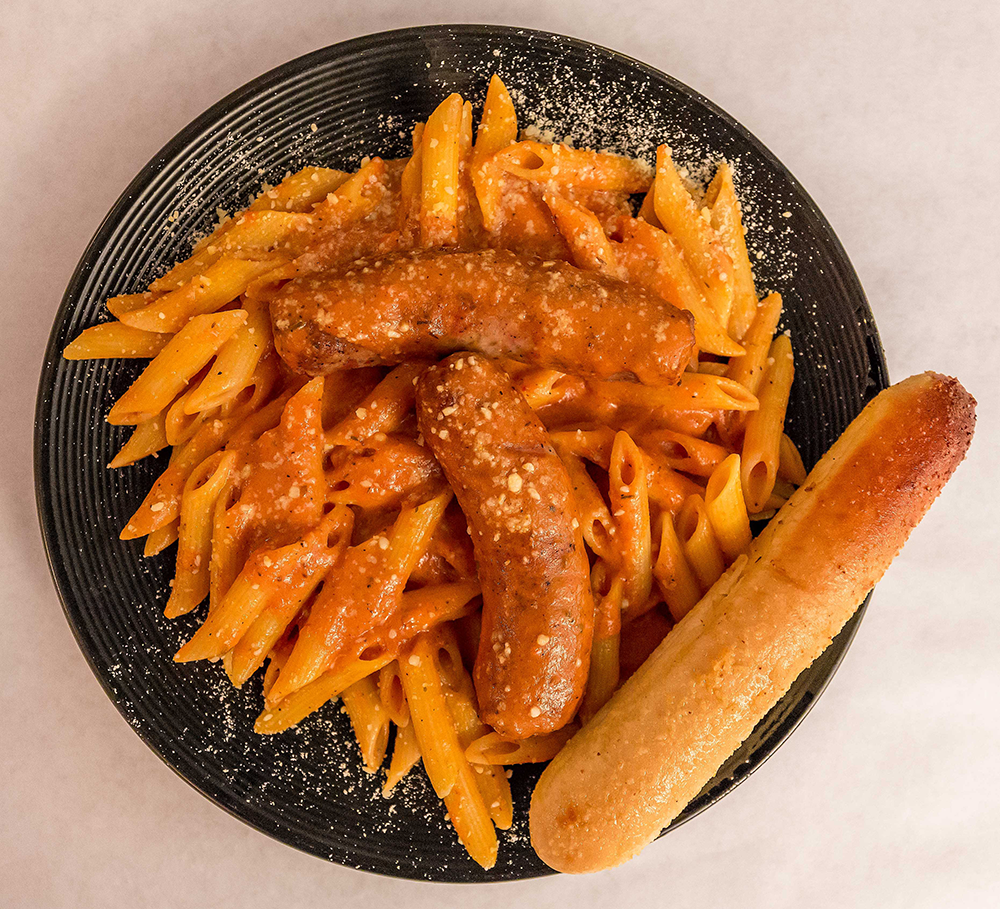 Penne pasta in tomato sauce with sausage pieces on a black plate, with a breadstick on the side
