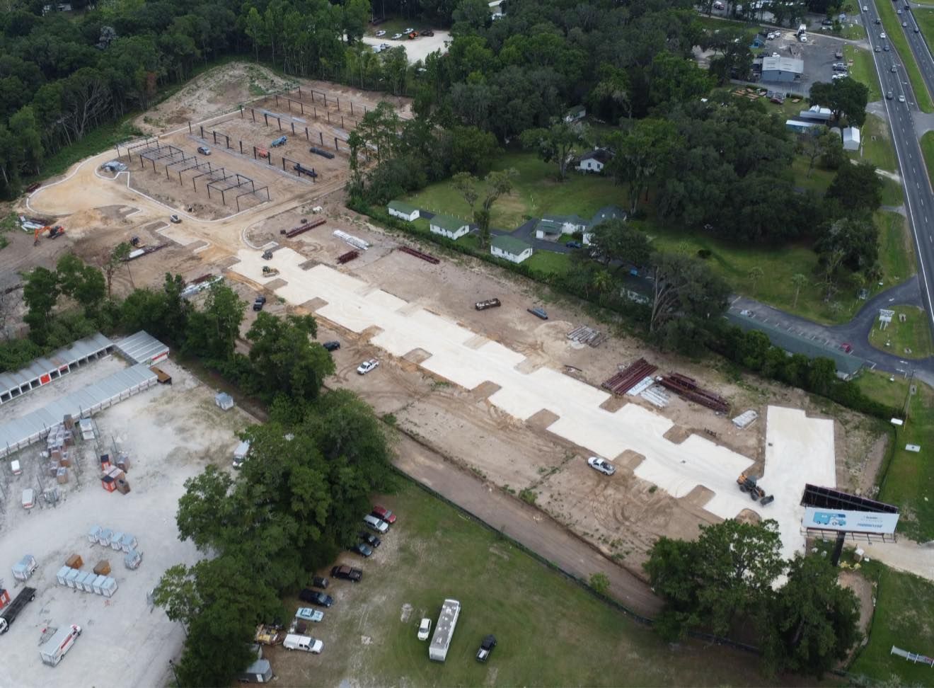 Aerial view of a large construction site with concrete foundations, dirt lots, trees, and a road nearby
