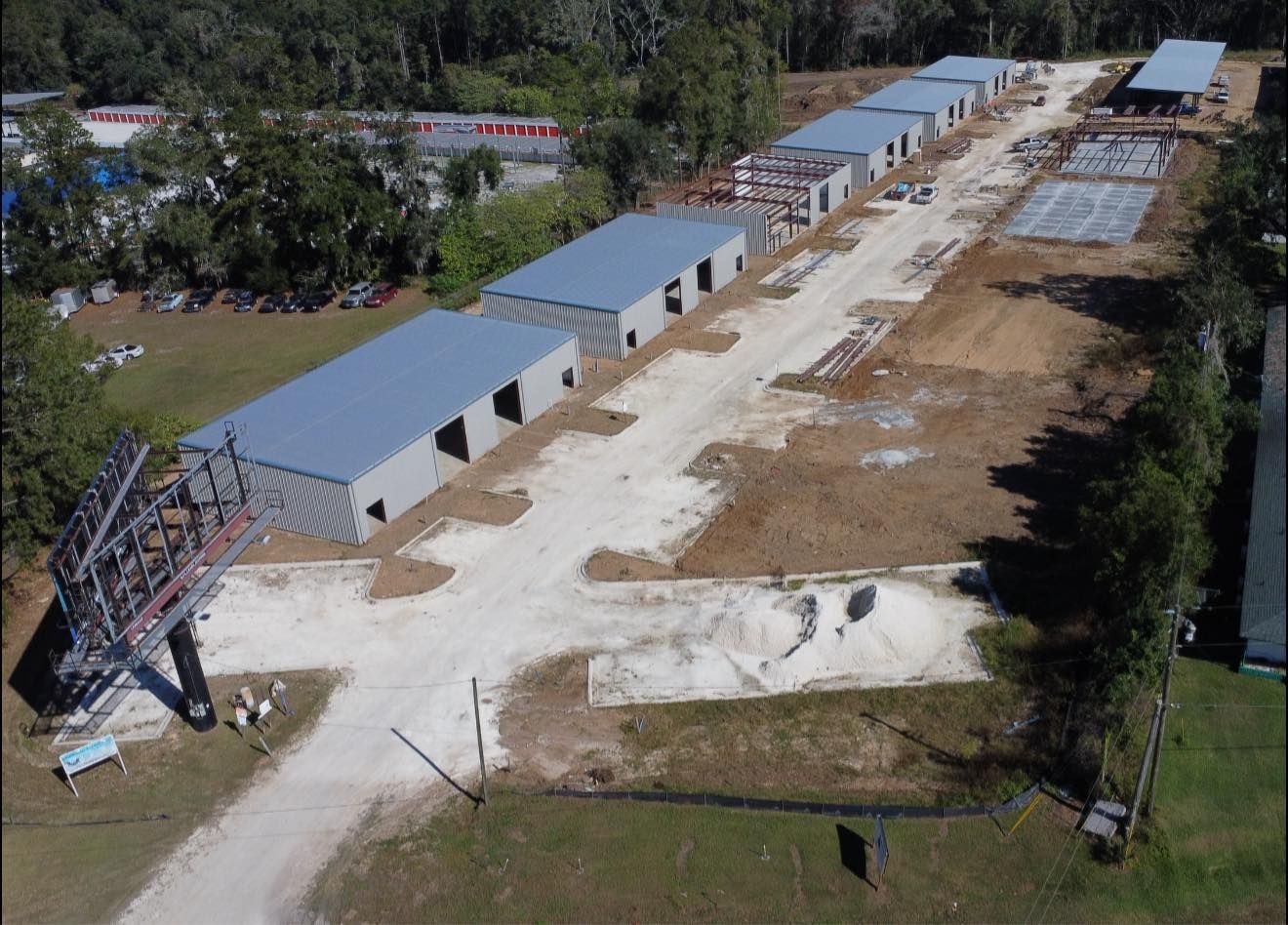 Aerial view of a construction site with long gray buildings, dirt roads, and equipment near trees