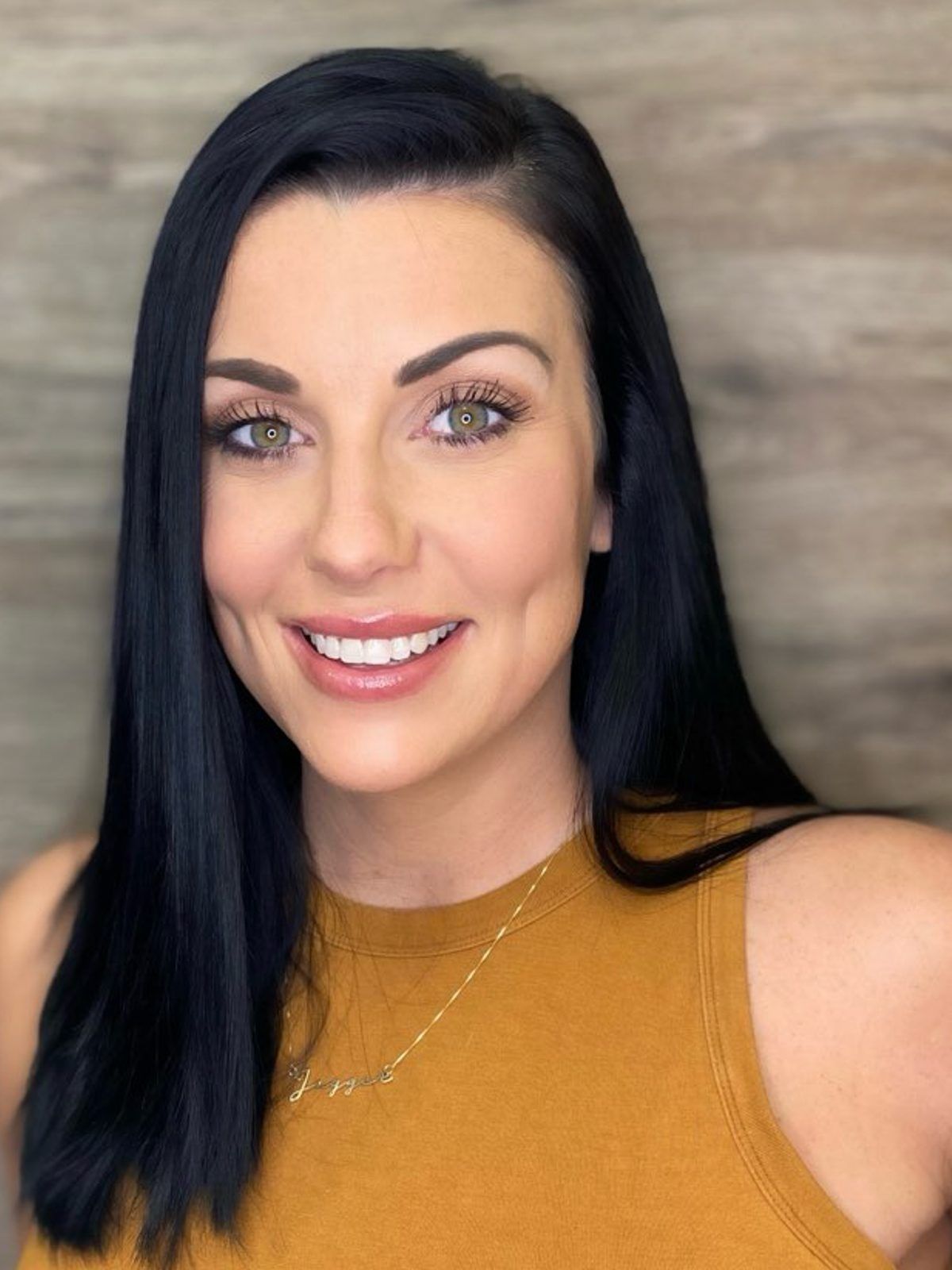 A person with long dark hair and a gold necklace smiles at the camera against a textured wooden background.