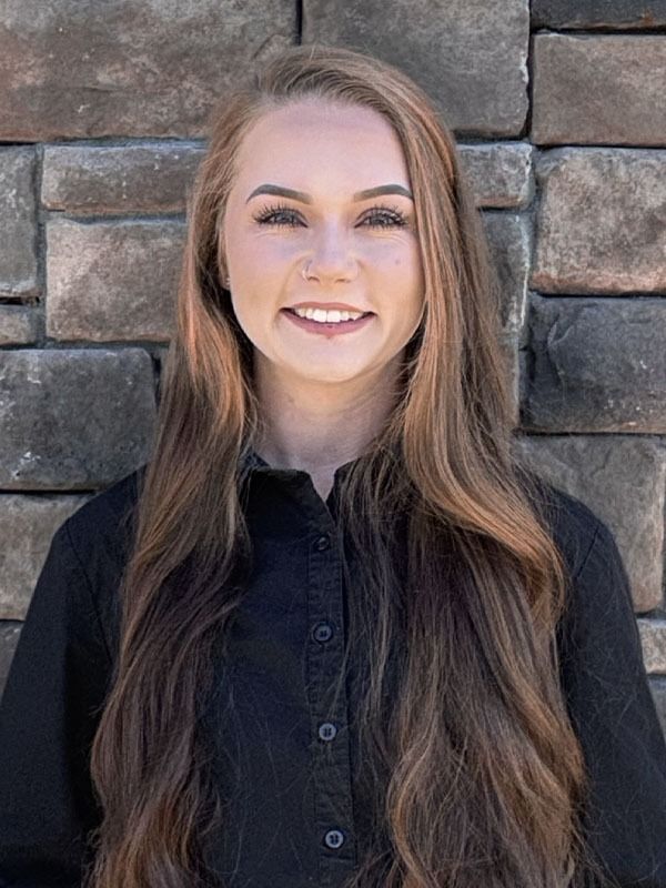 A person with long brown hair smiling at the camera, wearing a black collared shirt in front of a stone wall.