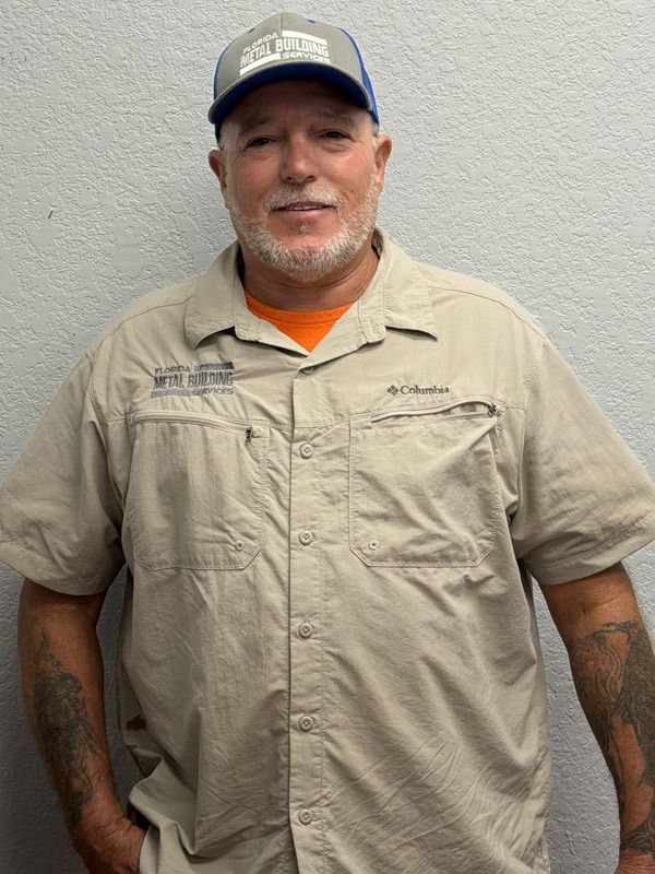 A person wearing a grey and blue baseball cap and a tan short-sleeved button-down shirt stands against a grey wall.