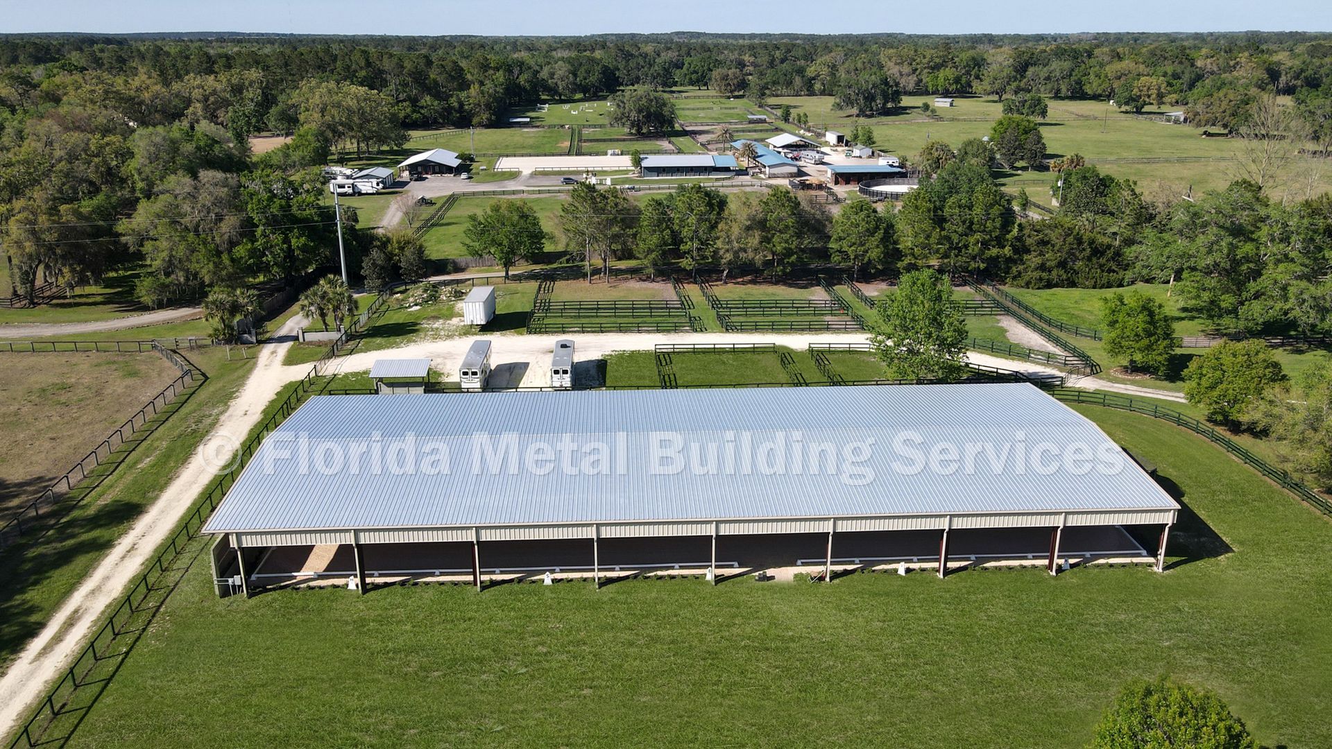 Aerial view of a large white-roofed barn in a green field with trees and buildings in the background