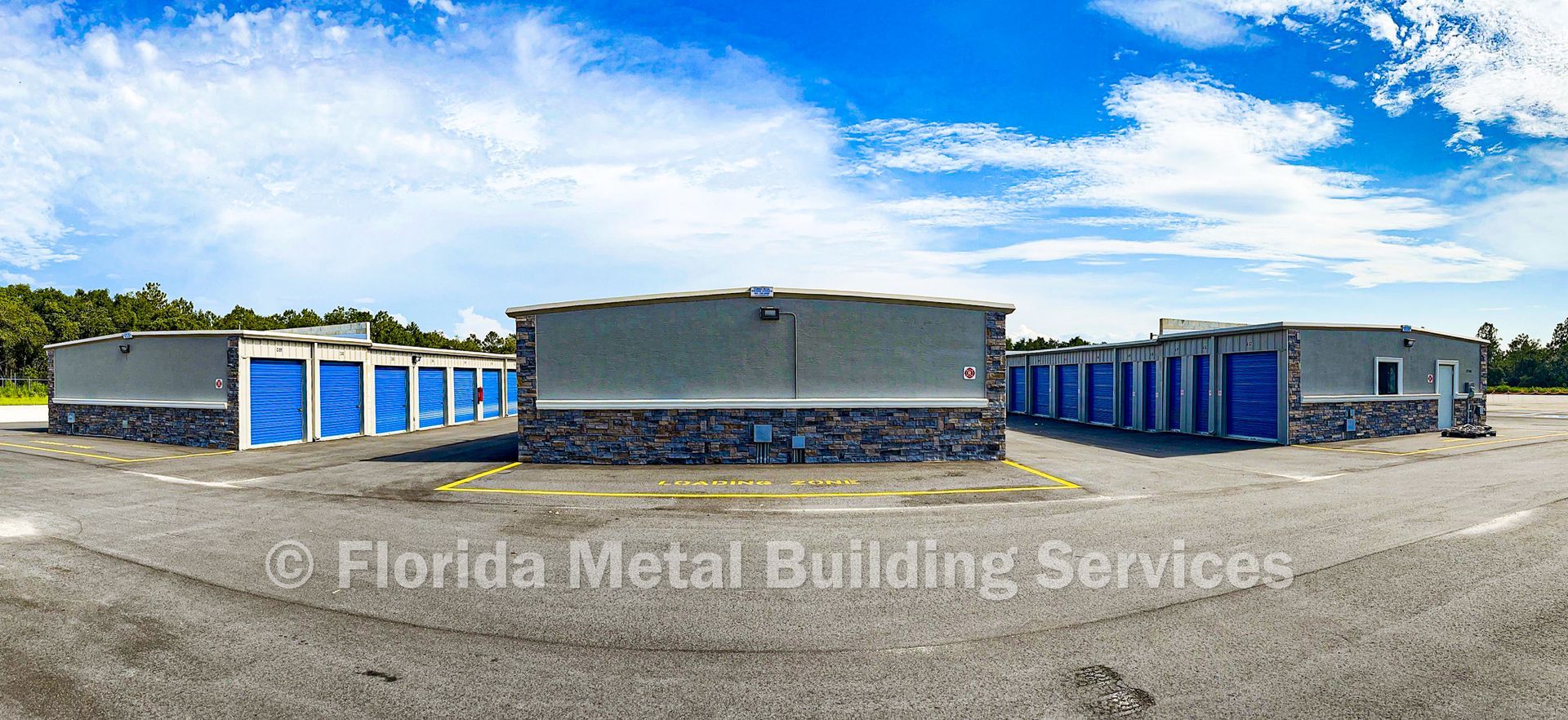 Wide-angle view of blue-and-gray metal buildings on a gravel lot under a partly cloudy sky