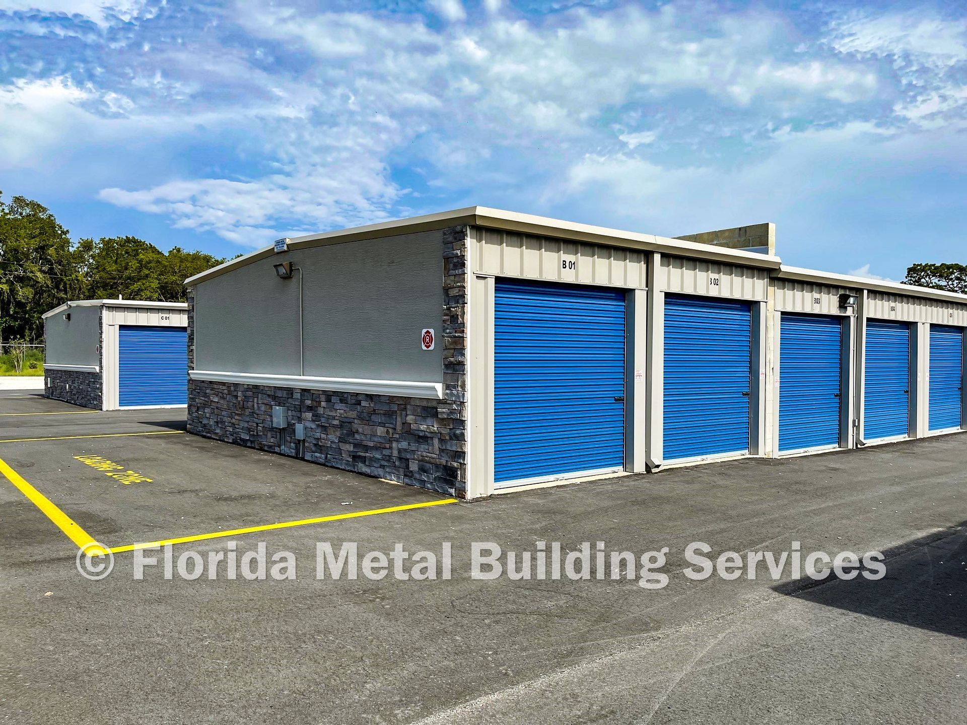 Blue roll-up storage units in a gray metal building with stone trim on an asphalt lot under a blue sky