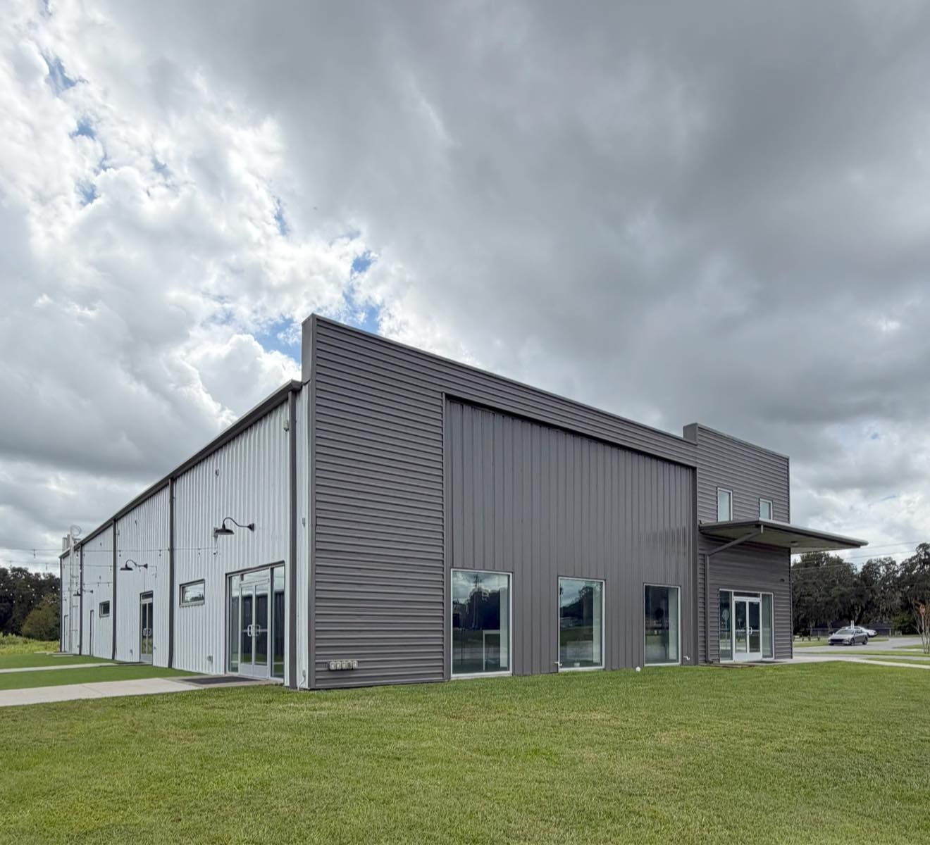 Modern gray metal warehouse building with large windows and a grassy foreground under cloudy skies