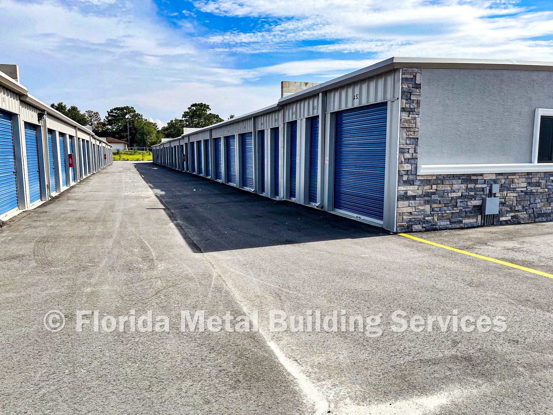 Storage facility with rows of blue roll-up doors along a gravel lane under a partly cloudy sky