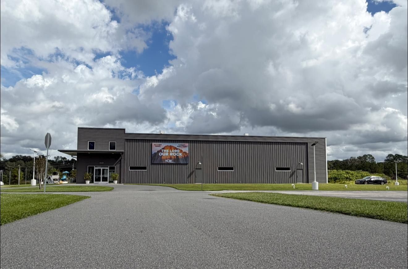 Modern gray building with a sign, seen from a wide gravel drive under cloudy skies