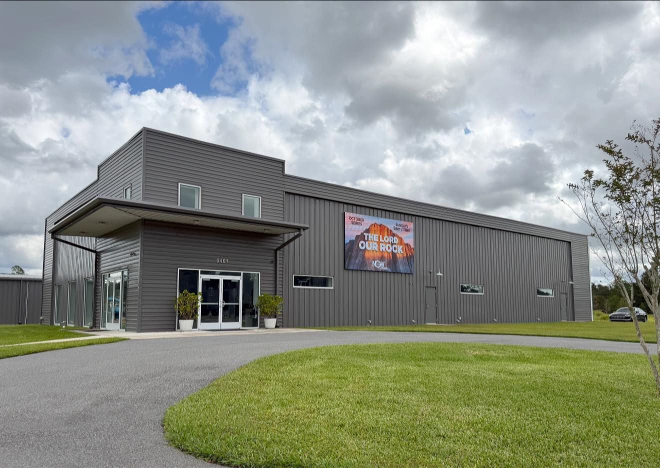 Modern gray building with glass entrance and a sign, viewed from a grassy parking area under cloudy skies