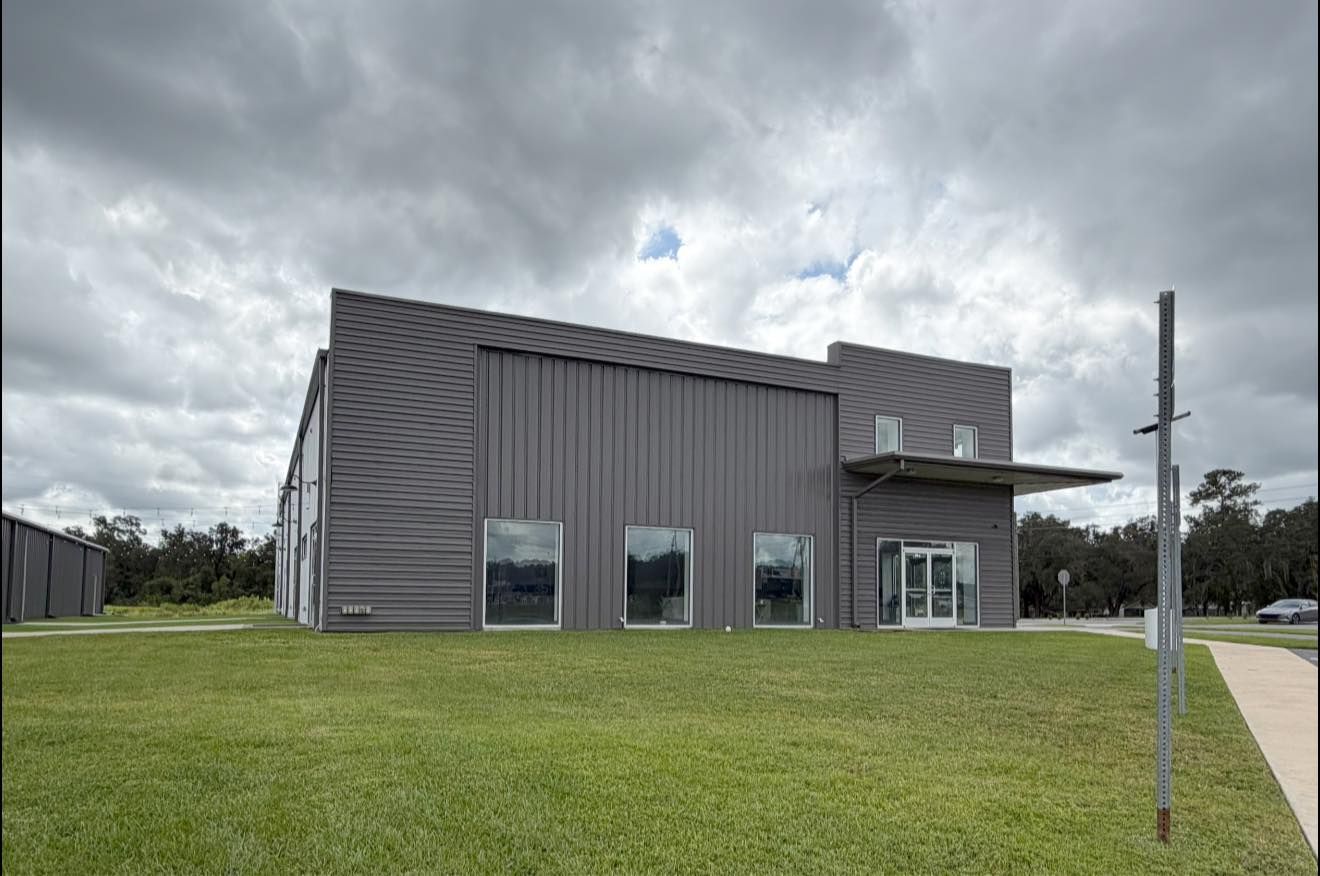 Modern gray metal building with tall windows and a small entry, set on a grassy lot under cloudy skies